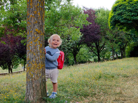 curly blond child with a briefcase on his back peeps out from behind a tree with a joyful smile on his faceの写真素材