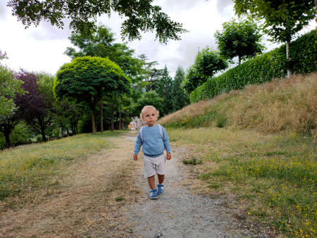 curly blond child with a briefcase behind his back runs along the path in the parkの写真素材