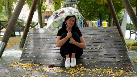 middle aged brunette woman sitting on a wooden surface with a baby umbrella in her hands in early autumnの写真素材