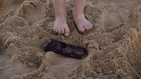 Phone thrown in the sand next to the feet of a childの写真素材