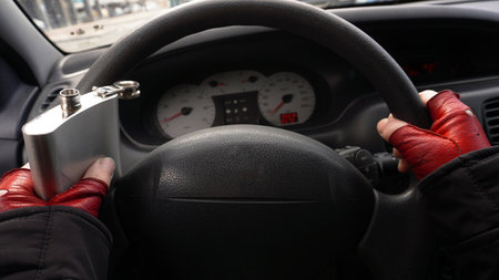 hands on the steering wheel of a car in red gloves, with a flask filled with alcohol in one hand, a person drives a car. Concept: Pyanstvo while driving a carの写真素材