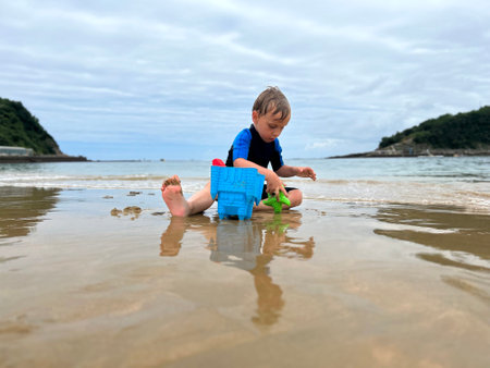 a blond kid with neoprene on. He is playing on the edge of the beach, with the sand using beach toysの写真素材
