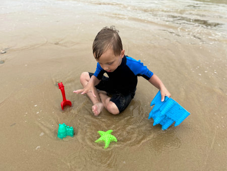 a blond kid with neoprene on. He is playing on the edge of the beach, with the sand using beach toysの写真素材