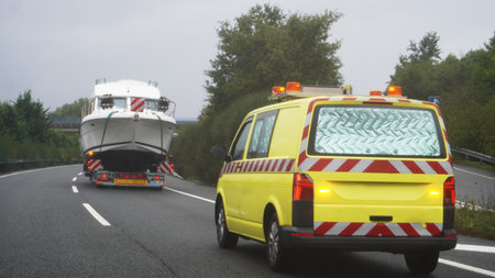 A truck with a large ship is driving along the road, accompanied by an escort car.