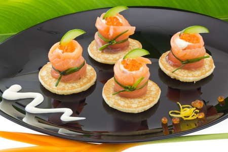 Red salmon roll with cheese, caviar and cucumber base, on black plate, with green leaf and tropical flowerの写真素材