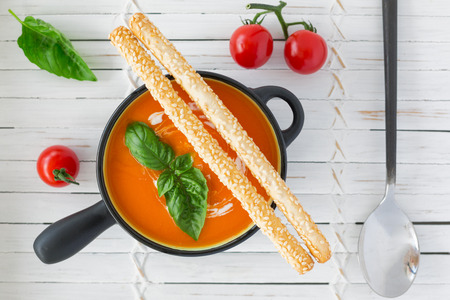 Overhead view of a Tomato soup in a black casserole, drizzled with cream, and garnish with leaves of fresh green basil and long bread sticks, on white wooden backgroundの写真素材