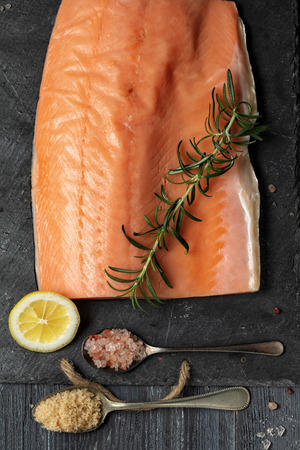 Raw salmon fillet on black slate plate and ingredients for making gravlax. Salt, sugar, lemon and rosemary in the background for dry cure marinade, dark background.の写真素材