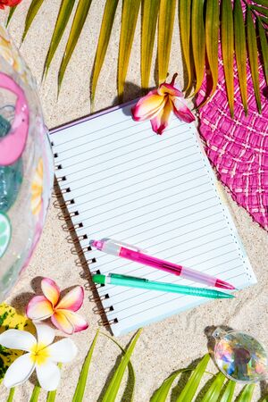 Open blank writing note pad with pink and green pen on sand, surrounded by green palm leafs, sea shells, pink hat, inflatable toys. Summer beach background, flat lay, vertical compositionの写真素材