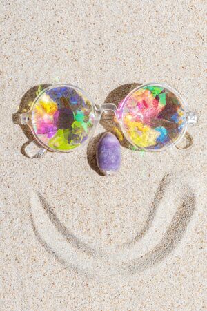 Funny smiling face made of kaleidoscope multicolor glasses and purple sea shell on sand background with copy space, flat lay, vertical compositionの写真素材