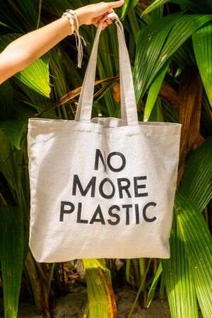 Female hand holding eco bag with sign No More Plastic on green tropical foliage background, vertical compositionの写真素材