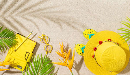 Yellow round hat, notepad and sunglasses with orange tropical flowers and palm leaf on sandy beach,...