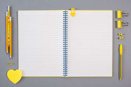 Assorted yellow office and school stationery supplies with open blank block note on gray background,...