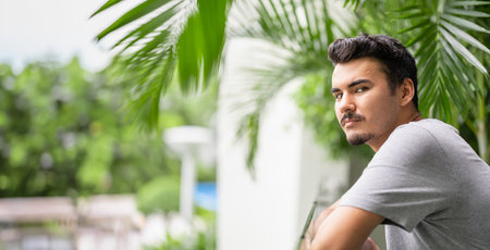 Young Man in gray shirt leaning on railing, lost in thought, with tropical plantsの写真素材