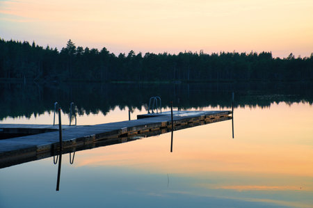 Wooden jetty at sunset on a Swedish lake. The colors of the sunset are reflected in the water. A bathing lake in the Scandinavian countrysideの写真素材