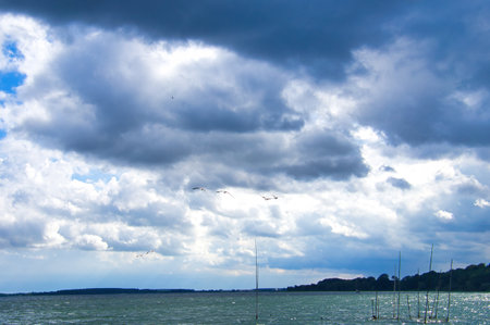Stormy sky over a lake. Dark clouds contrast with rays of sunlight that make the water sparkle. Four birds fly in the foreground.の写真素材