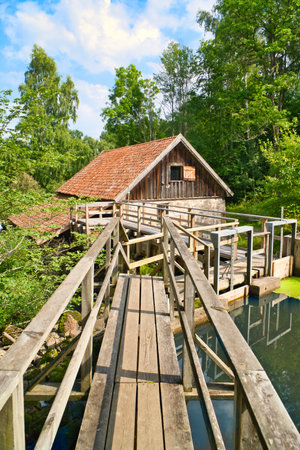 Old watermill surrounded by green nature. A small stream flows over moss-covered stones, surrounded by trees. Idyllic landscape with rustic wooden architecture.の写真素材