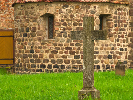 A historic stone cross in front of an old rustic church with small windows. Lively green grass in the foreground adds depth and contrast to the picture.の写真素材
