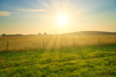 Sunset over a wide field with rolling hills in the background, a wooden fence in the foreground. Warm mood lighting and silhouettes emphasize the landscape.の写真素材
