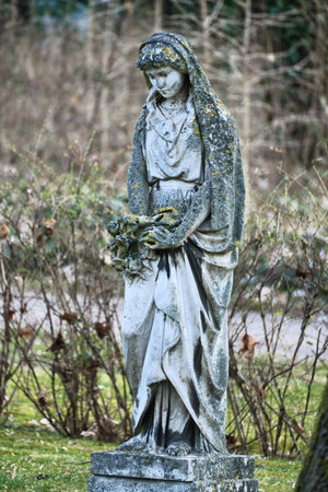 Moss-covered female statue with headscarf, surrounded by wild plants. The elegant sculpture holds flowers and stands in an enchanted garden. Mystical atmosphere.の写真素材