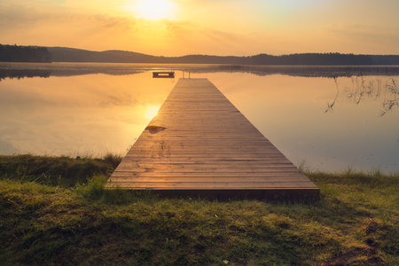 A wooden pier on a lake in the golden light of the sunset. The calm water surface, with rolling hills in the background, symbolizes peace, tranquility, and relaxation.の写真素材