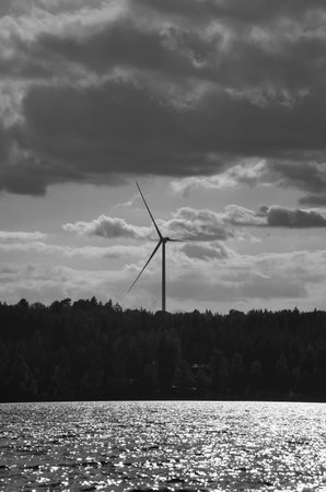A wind turbine rises behind a green forest on the lake shore. Sunlight reflects off the water,...