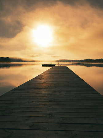 A wooden walkway leads into a tranquil lake, surrounded by mist and sunlight. A romantic morning mood with gentle reflections and a peaceful atmosphere.の写真素材