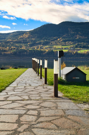Stone path with lighting, leads to the lake shore in front of picturesque mountains and blue sky, sunny autumn mood and peaceful landscape in Norway.の写真素材