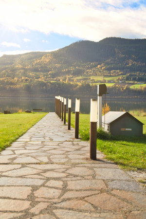 Stone path with lighting, leads to the lake shore in front of picturesque mountains and blue sky, sunny autumn mood and peaceful landscape in Norway.の写真素材