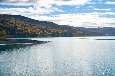 Clear water of a mountain lake, surrounded by autumn-colored hills under a blue sky with light clouds, a peaceful natural idyll.の写真素材