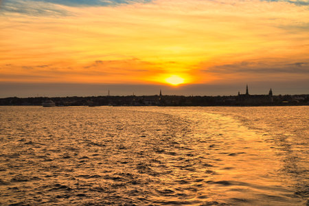 Romantic sunset over calm sea, gentle waves and boat wake in the evening light, silhouette of a coastal town on the horizon, maritime evening atmosphere.の写真素材