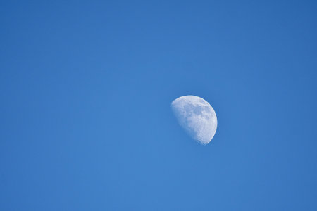 A clear blue sky with a clearly visible waxing moon, whose bright surface and fine structures stood out distinctly, creating a calm, minimalist scene.の写真素材