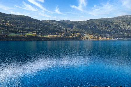 Clear water of a mountain lake, surrounded by autumn-colored hills under a blue sky with light clouds, a peaceful natural idyll.の写真素材