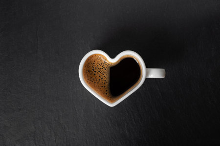 white cup in the shape of a heart with coffee and beautiful foam on a black stone background. view from above. copy spaceの写真素材