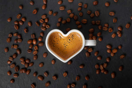 white cup in the shape of a heart with coffee and beautiful foam on a black stone background with scattered coffee beans. view from above. copy spaceの写真素材