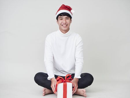 Christmas concept. Young man celebrate merry christmas and happy new year wear santa hat in hand holding a gift box tied with red ribbons on a white background.の写真素材