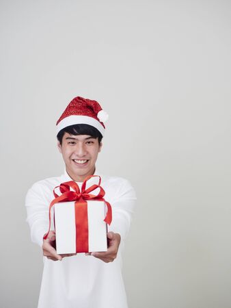 Christmas concept. Young man celebrate merry christmas and happy new year wear santa hat in hand holding a gift box tied with red ribbons on a white background.の写真素材
