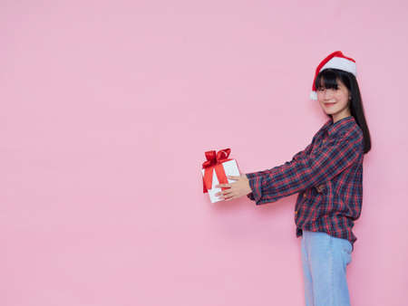 Cheerful young girl wearing santa hat giving gift box isolated over pink background, space for textの写真素材