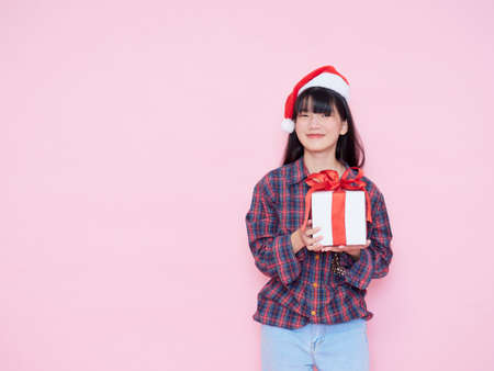 Cheerful young girl wearing santa hat with holding gift box on pink backgroundの写真素材