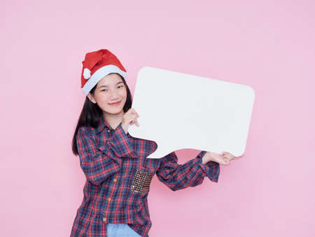 Cute teenage gir in santa hat pointing finger on blank poster while standing on pink background.