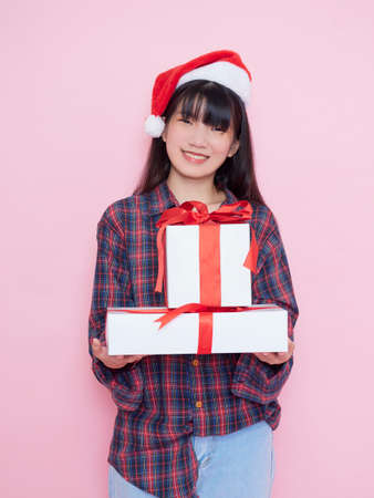 Cheerful young girl wearing santa hat with holding gift boxes on pink backgroundの写真素材