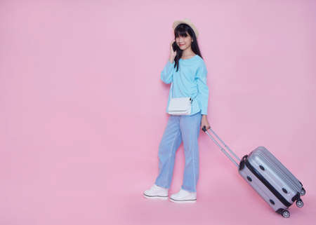 Young woman with suitcase and talking on phone on pink background, space for text. Summer vacation.