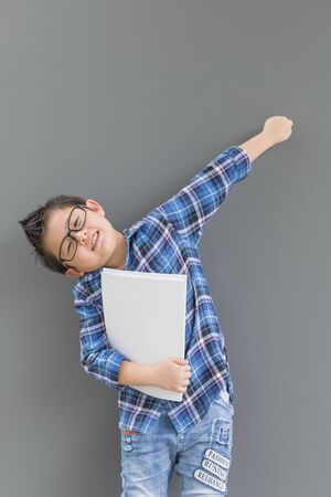 Asian boy raise hand up feels happiness when time to school and his hand hold the book against the gray wallの写真素材