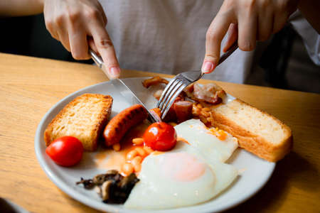 a girl with a knife and fork eats an English breakfast fresh tomatoes scrambled eggs fried eggs with rye white fresh bread and meat sausage pork sausage meat food tomorrow lunch meal healthの写真素材
