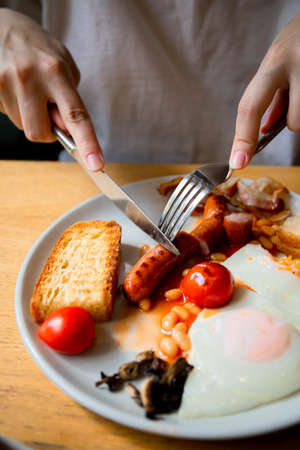 a girl with a knife and fork eats an English breakfast fresh tomatoes scrambled eggs fried eggs with rye white fresh bread and meat sausage pork sausage meat food tomorrow lunch meal healthの写真素材