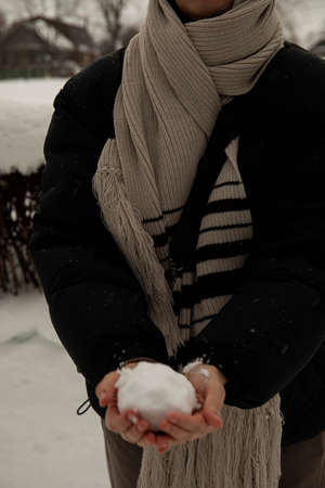 a girl in a black jacket and a gray scarf holds a snowball in her hands, crumples it and throws it....