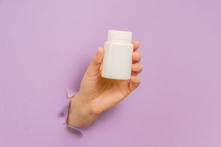 View on white bottle plastic tube in hands on a lilac background. Packaging for pills, capsules, supplements. Cosmeticsの写真素材