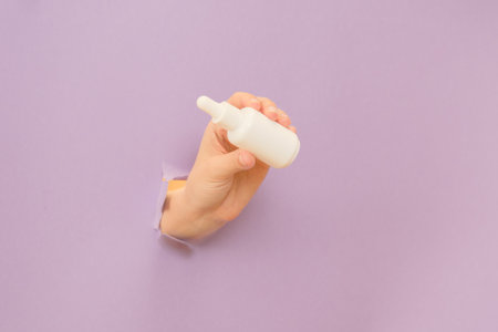 Hand holding glass bottle with dropper lid. White container with cosmetic product, serum (essential oil ) on a lilac background. Concept of beautyの写真素材
