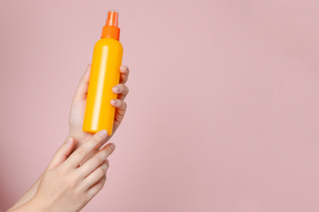 Orange spray cosmetic bottle in female hand on the solid pink background. Package, your brand