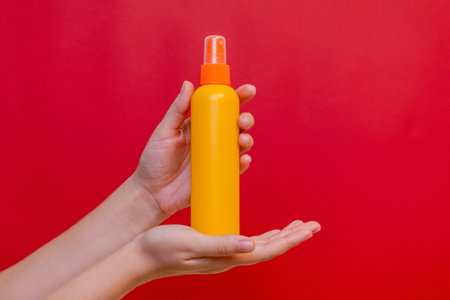 Female hand holding a orange spray cosmetic bottle on the solid red background. Package, your brandの写真素材