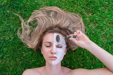 Woman's face with cosmetic face mask and with roller smoothing wrinkles in the grass. Spa, healthy,...